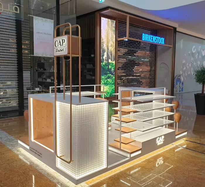Retail kiosk with illuminated display units and a branded sign on a tall frame inside a shopping centre