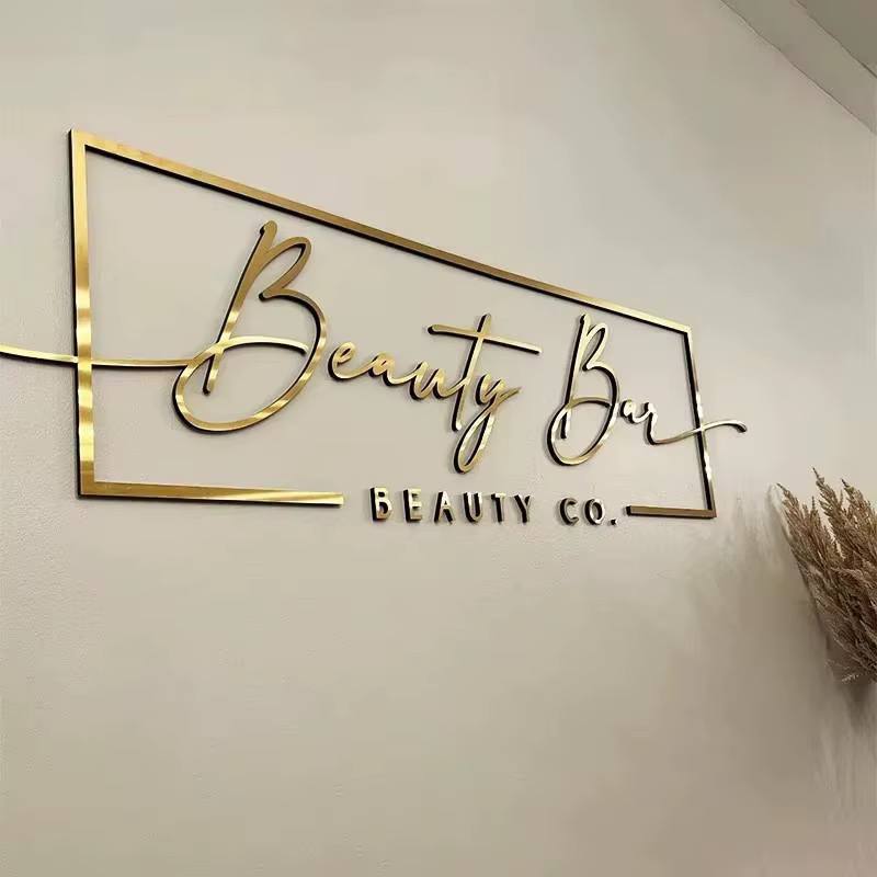Gold mirror script logo inside a thin gold outline frame with small black 3D letters on a beige wall
