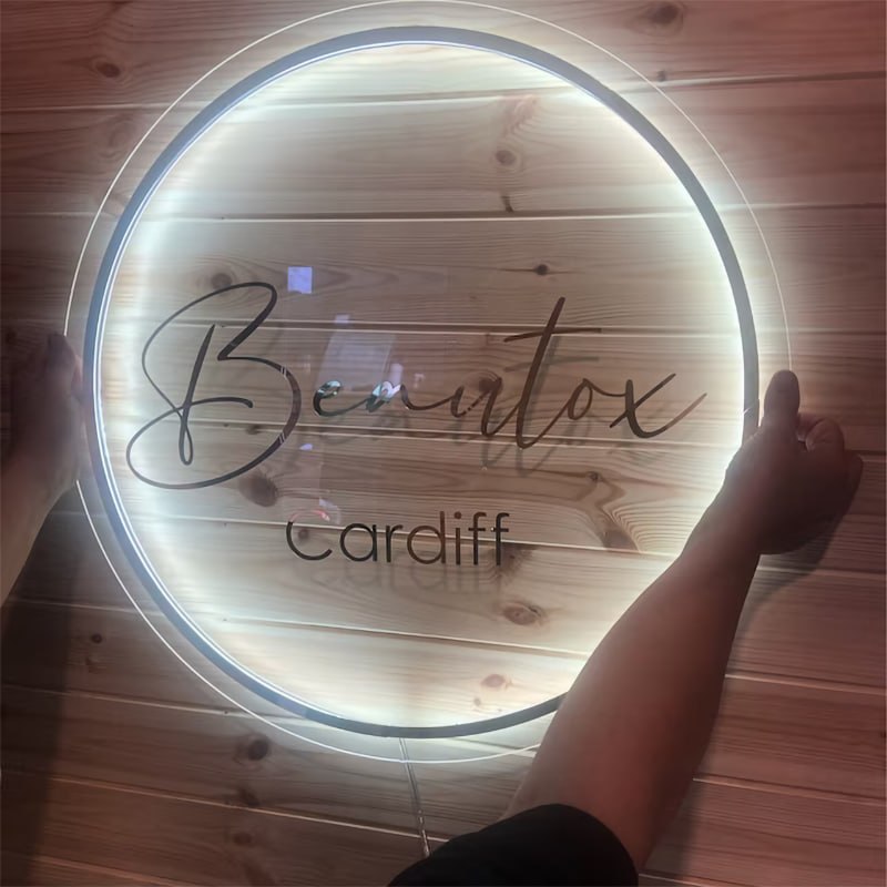 Backlit round clear acrylic wall sign with script logo text, held up to show the warm LED glow around the edge